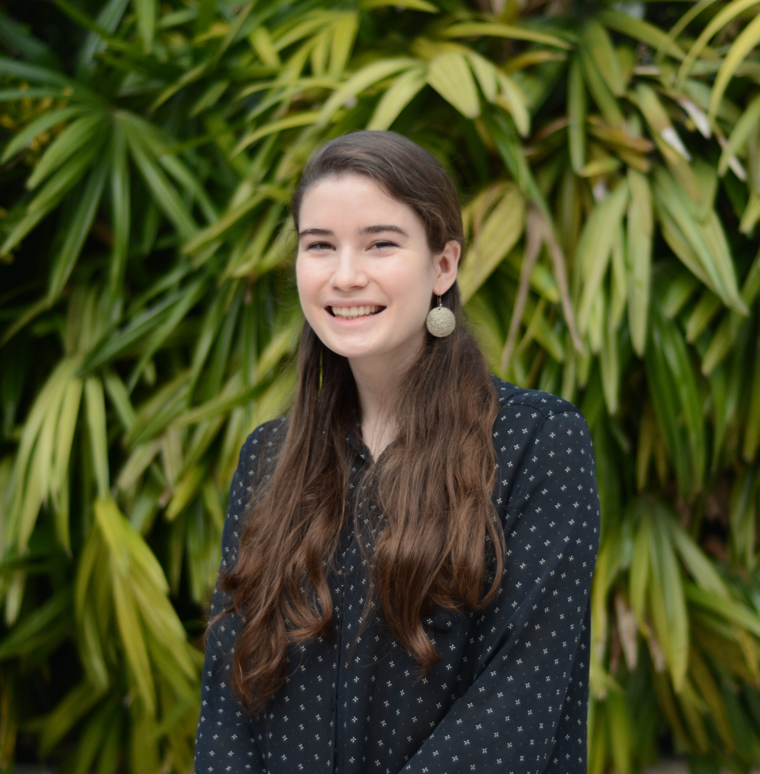 A white woman smiling with long dark hair by a backdrop of greenery