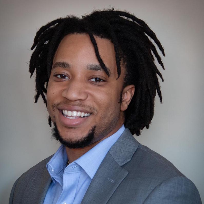 A black man in a suit with a blue shirt smiling before a grey backdrop