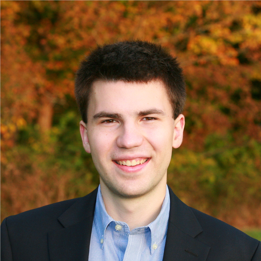 A white man with short black hair smiling before colorful fall foliage