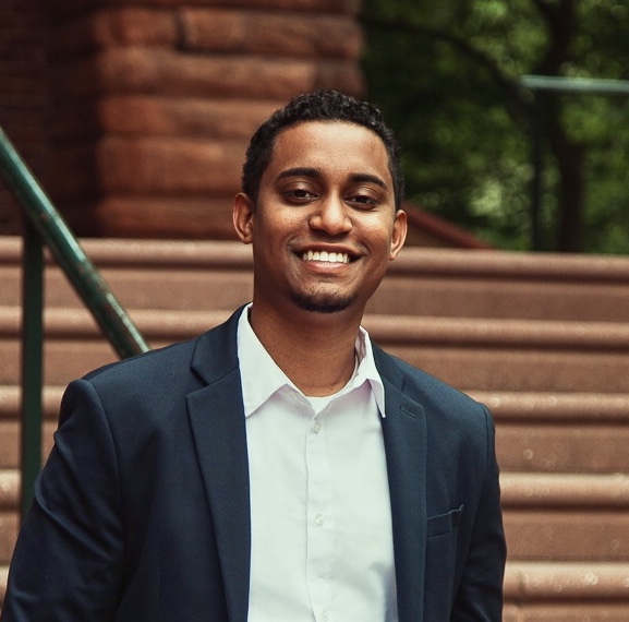 A black man smiling before a brick building with stairs