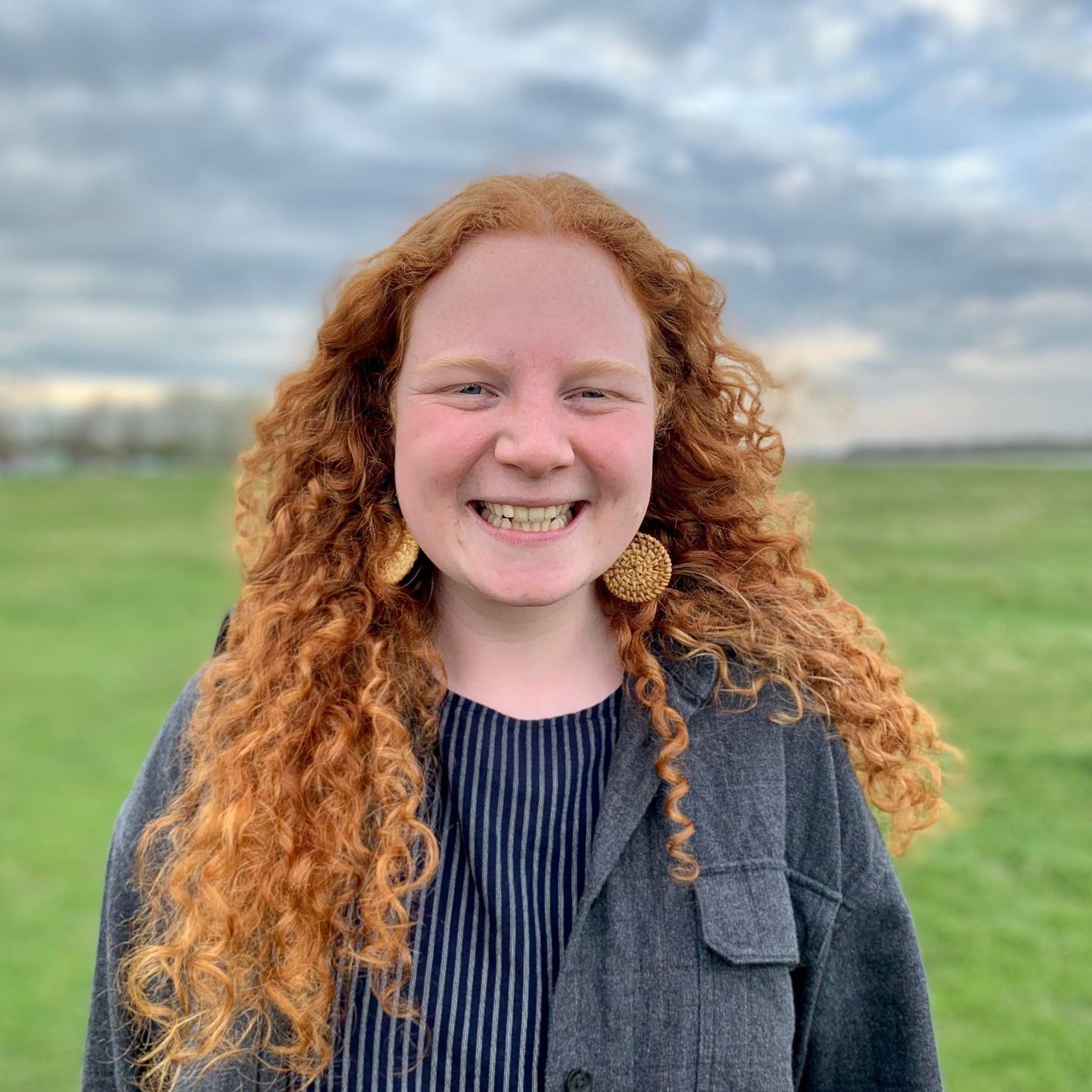 A woman smiling with long red curly hair with a grey striped sweater standing in a grassy field