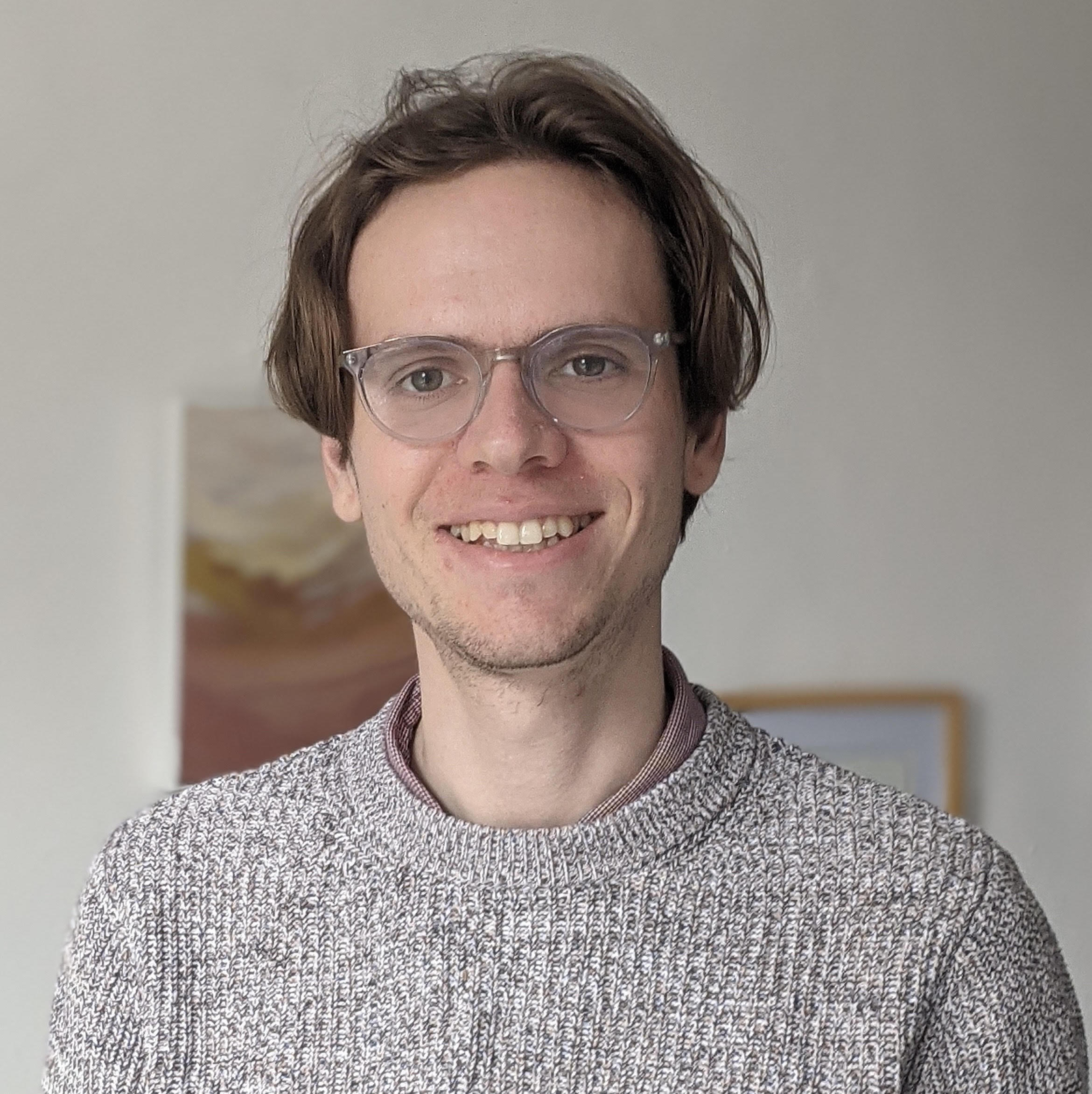 A man smiling with messy brown hair wearing glasses and a grey sweater