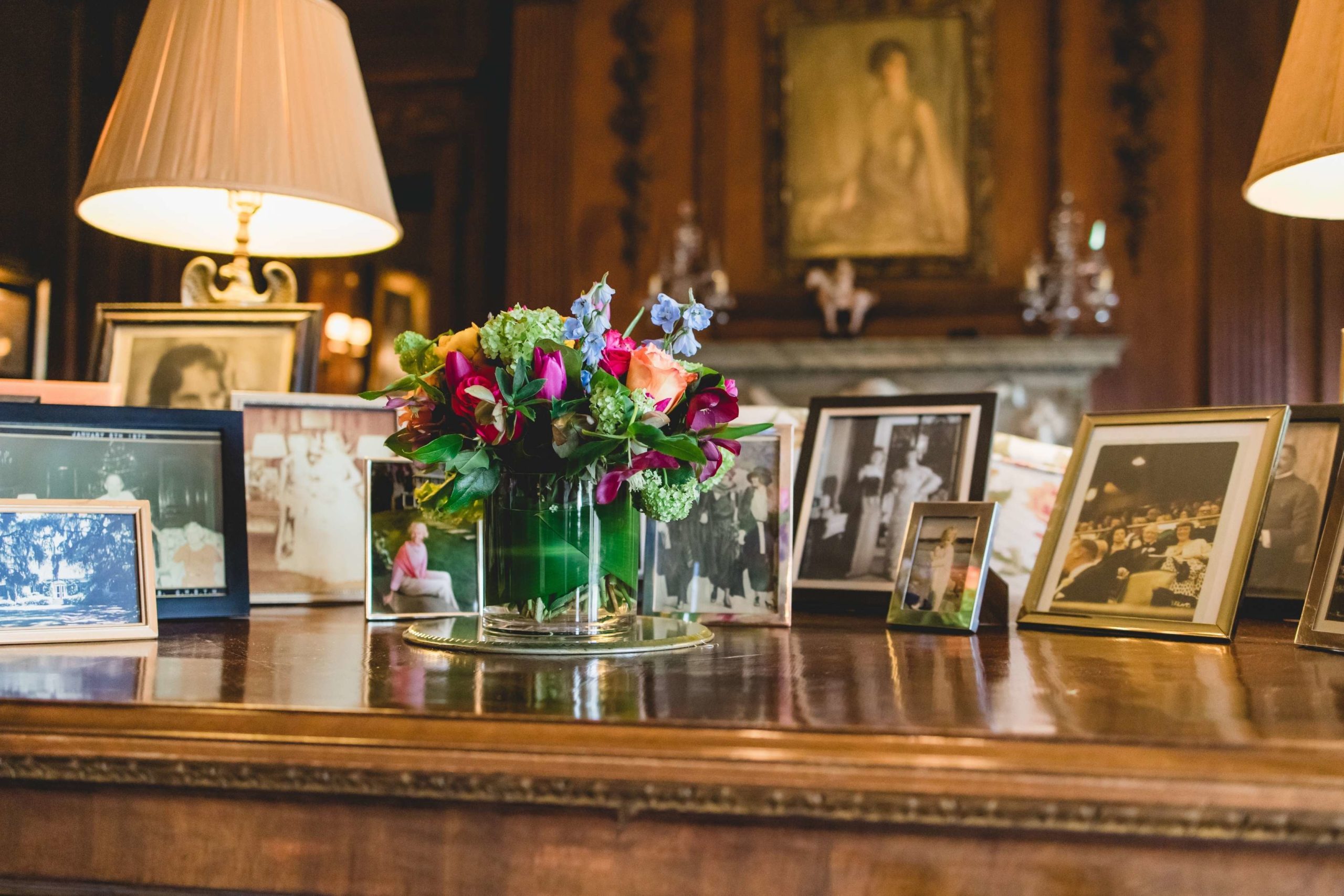 A vase of colorful flowers on a wooden table with many photographs in picture frames and two lamps