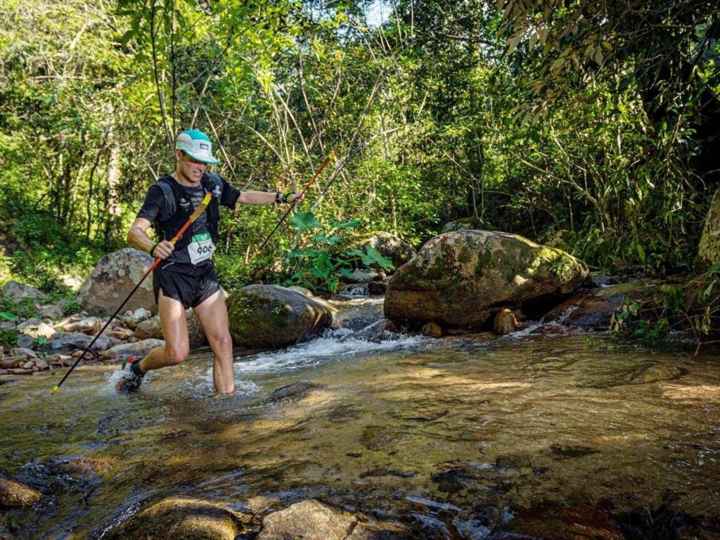 Sam Bennett, a white man, wearing a black tshirt, shorts and a baseball cap uses walking sticks as he crosses a stream during a cross country hike