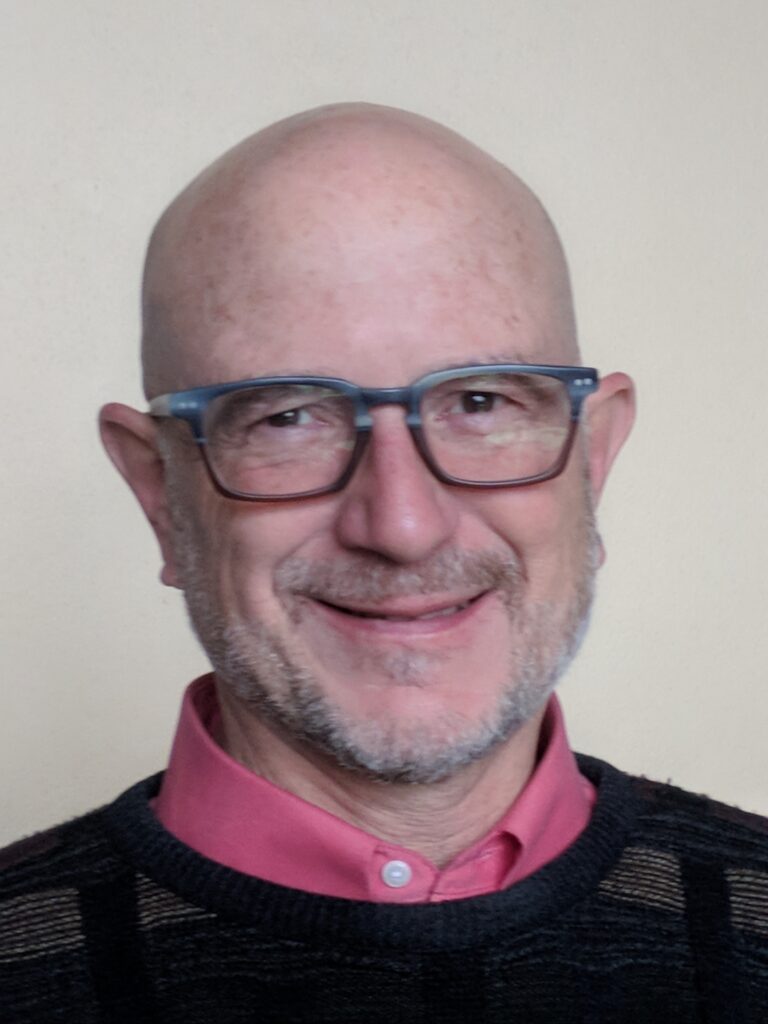 Brooks Kolb, a white man with glasses wears a button up under a sweater in front of a white wall