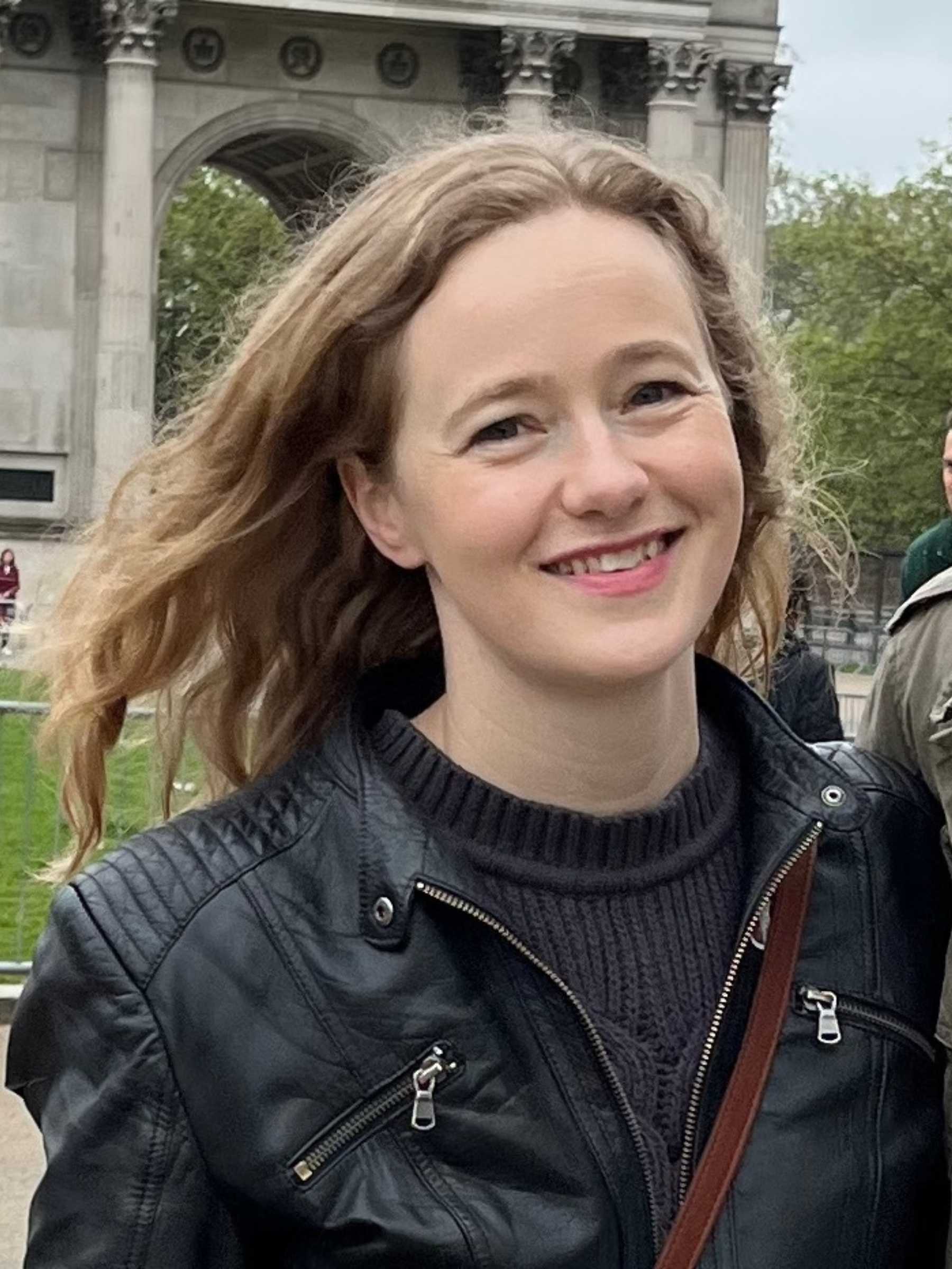 Ruth stands in a London park wearing a black sweater and a black leather coat